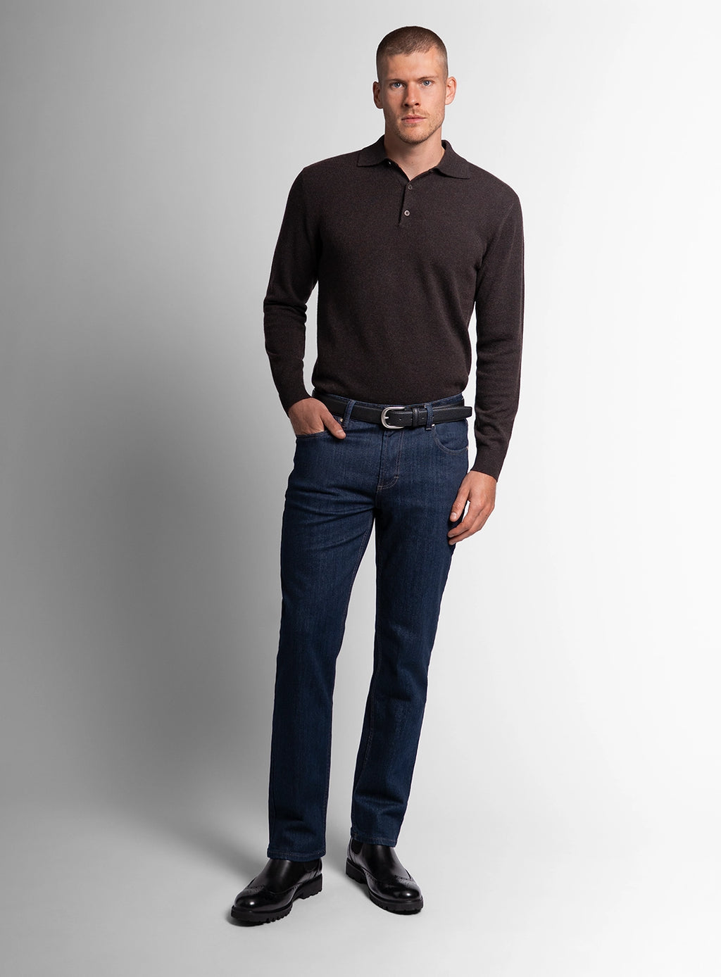 Man wearing a dark brown long-sleeve polo shirt and blue jeans on a gray background