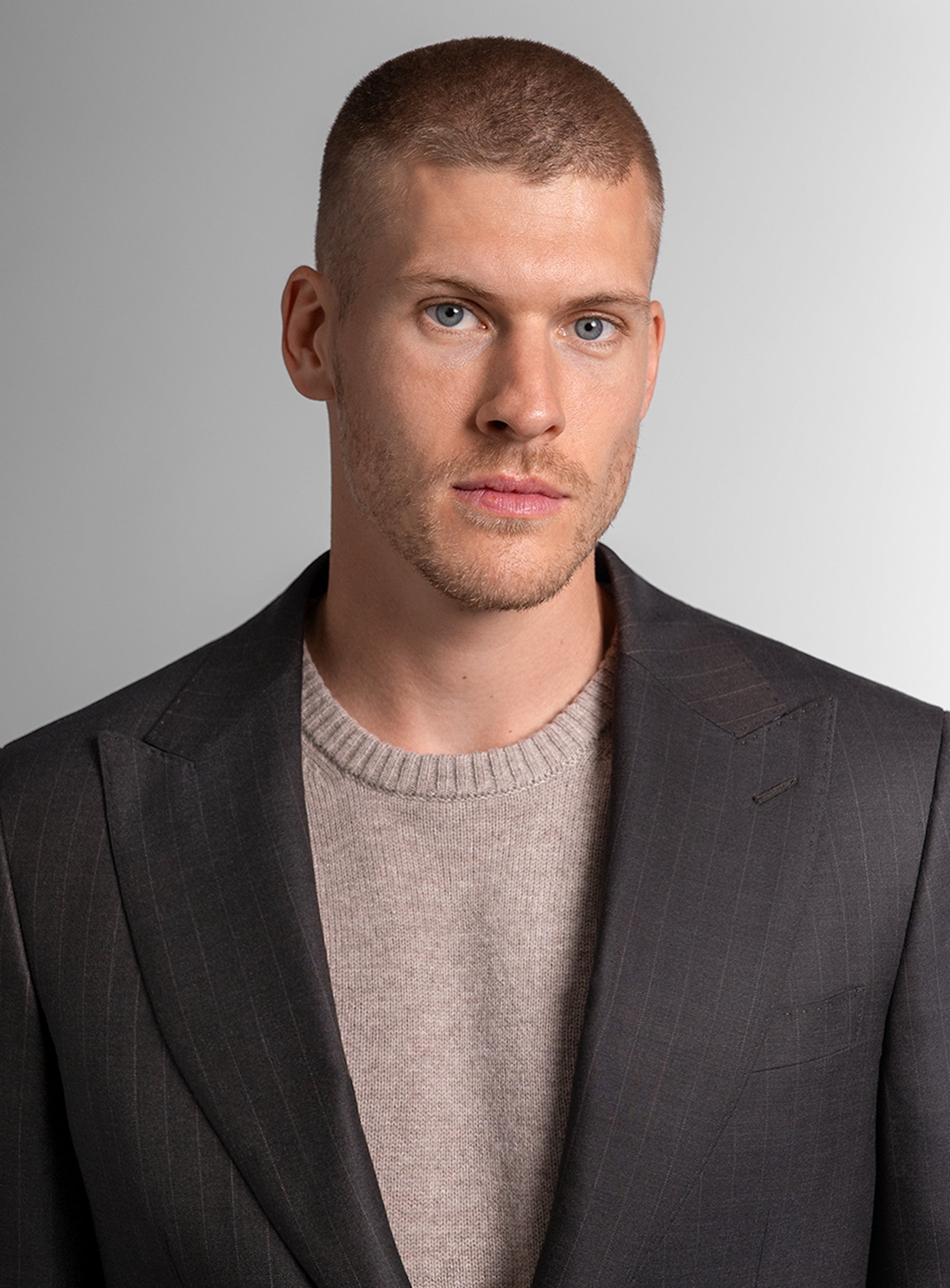 Man wearing a brown suit jacket over a light gray sweater against a plain background