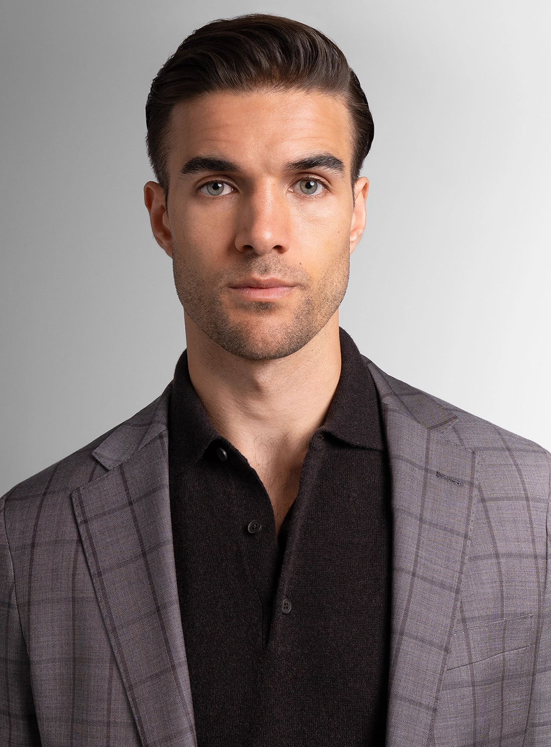 Man wearing a gray checkered suit jacket over a black shirt on a plain background