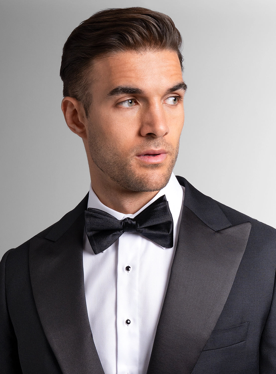 Man wearing a black tuxedo with a bow tie against a gray background