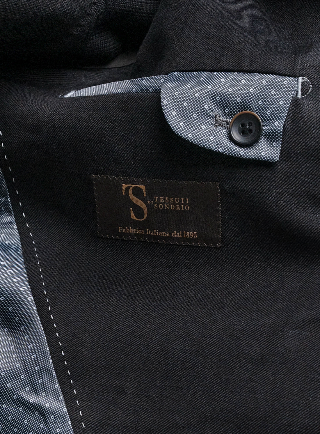 Close-up of a black garment with a Tessuti Sondrio label, featuring a button and pocket detail.