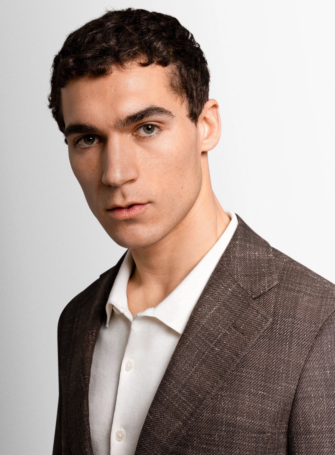 Man wearing a brown suit jacket over a white shirt on a light gray background
