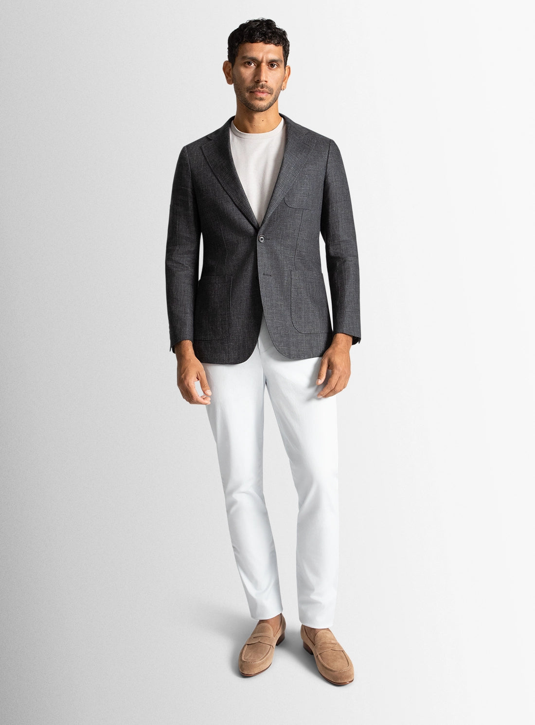 Man wearing a gray blazer, white shirt, white pants, and brown shoes on a light gray background