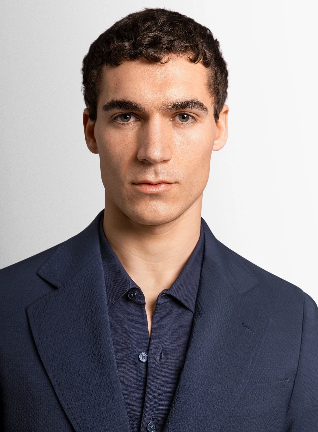 Man wearing a navy blue suit jacket over a dark shirt against a light gray background