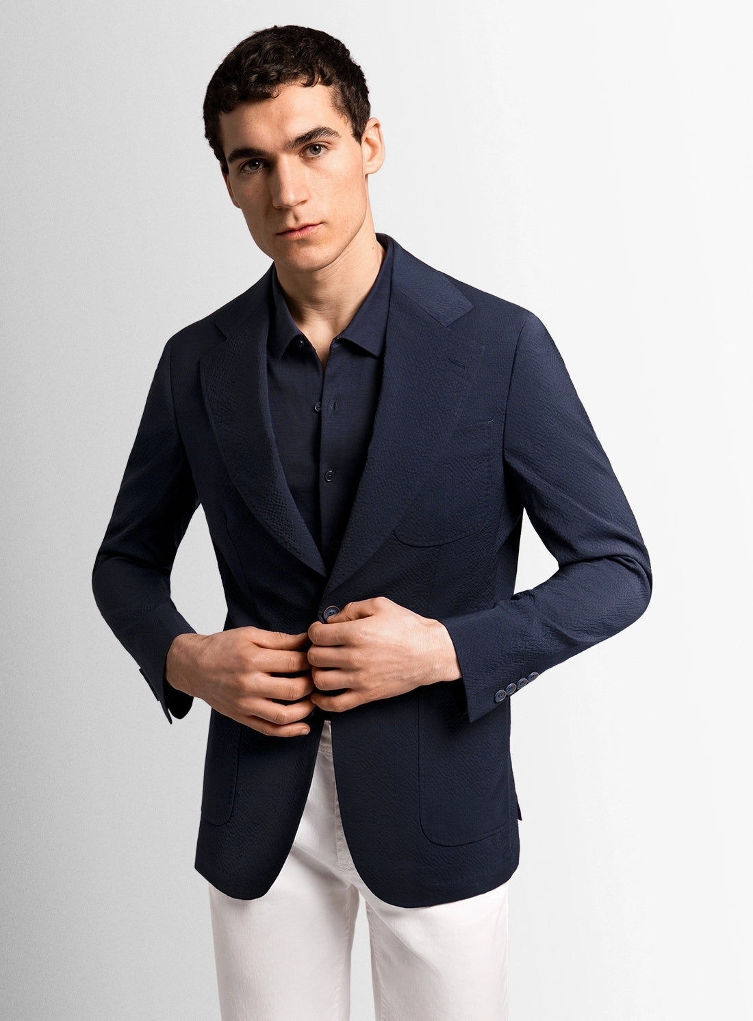Man wearing a navy blazer over a black shirt with white pants on a light gray background