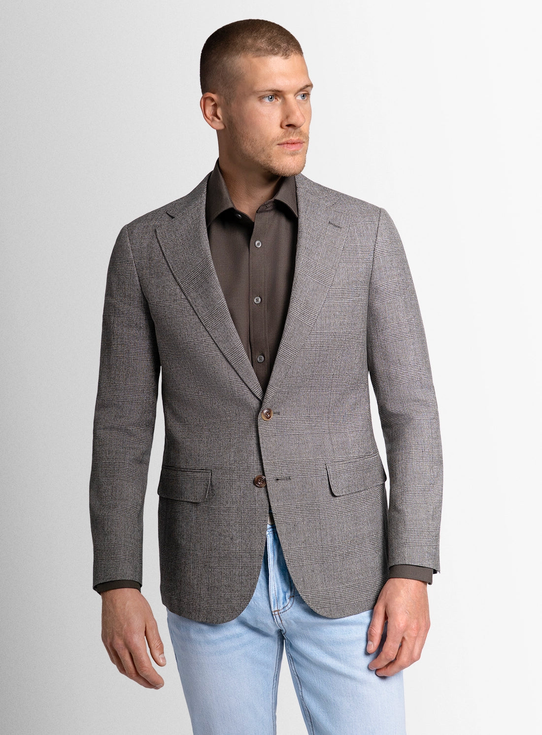 Man wearing a gray blazer over a brown shirt with light blue jeans on a plain background