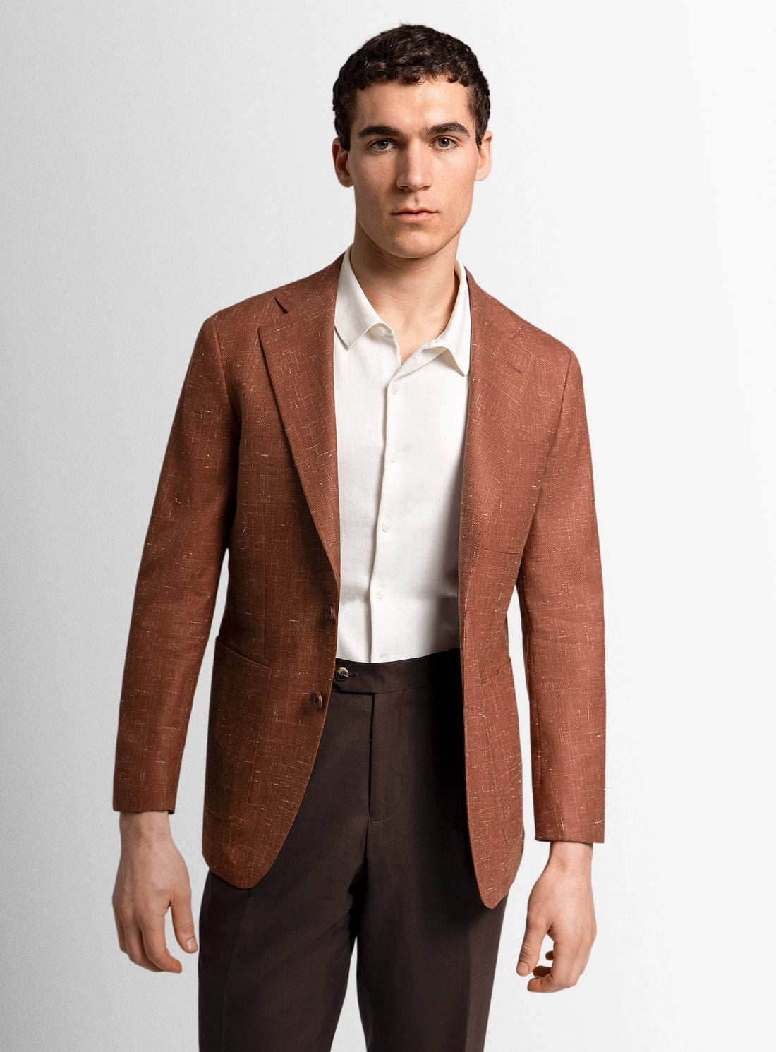 Man wearing a brown blazer over a white shirt and dark pants on a light gray background