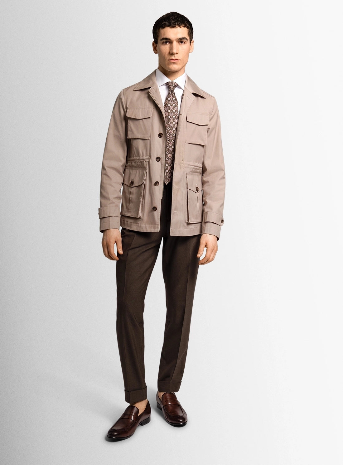 Man wearing a beige jacket, patterned vest, and brown pants on a white background