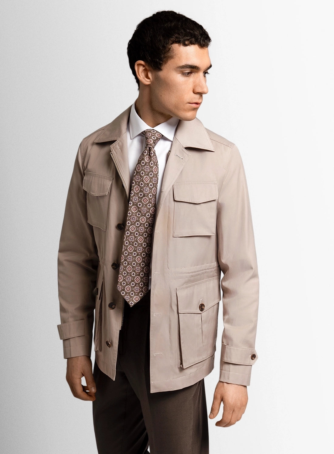 Man wearing a beige trench coat over a suit with a patterned tie on a plain background