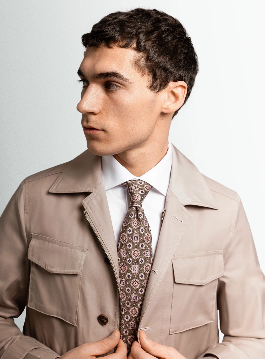 Man wearing a beige trench coat and patterned tie against a plain background