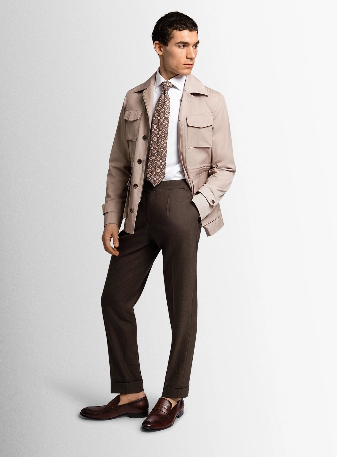 Man wearing a beige jacket, patterned tie, white shirt, and brown pants on a light gray background
