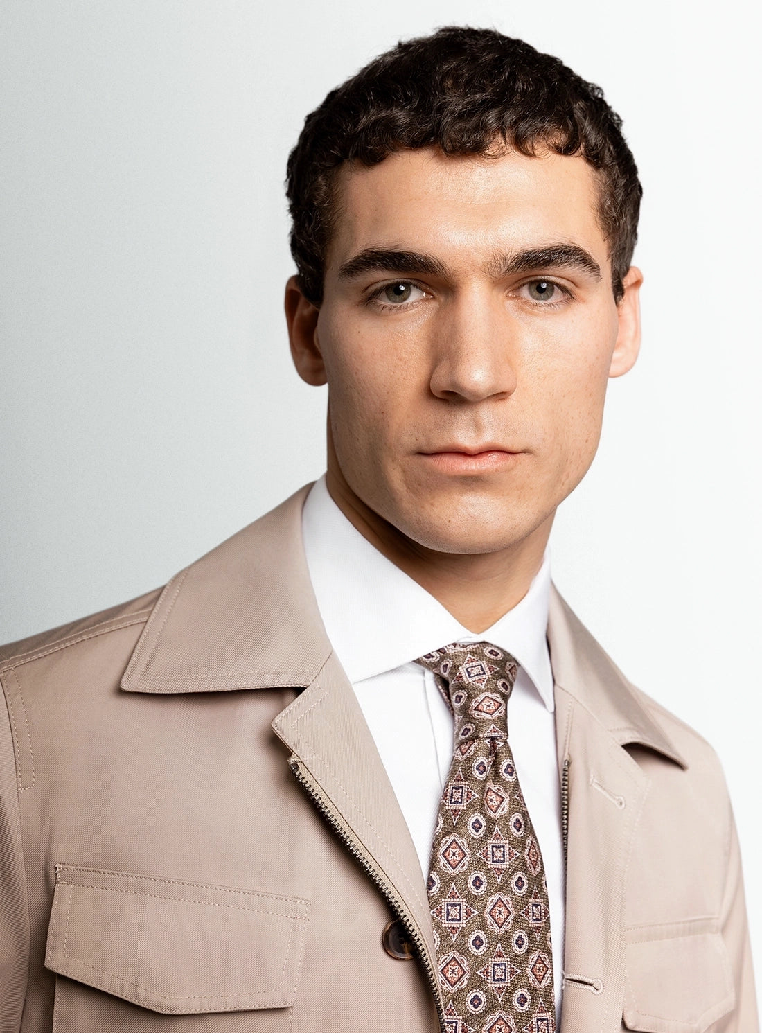Man wearing a beige trench coat and patterned tie against a plain background