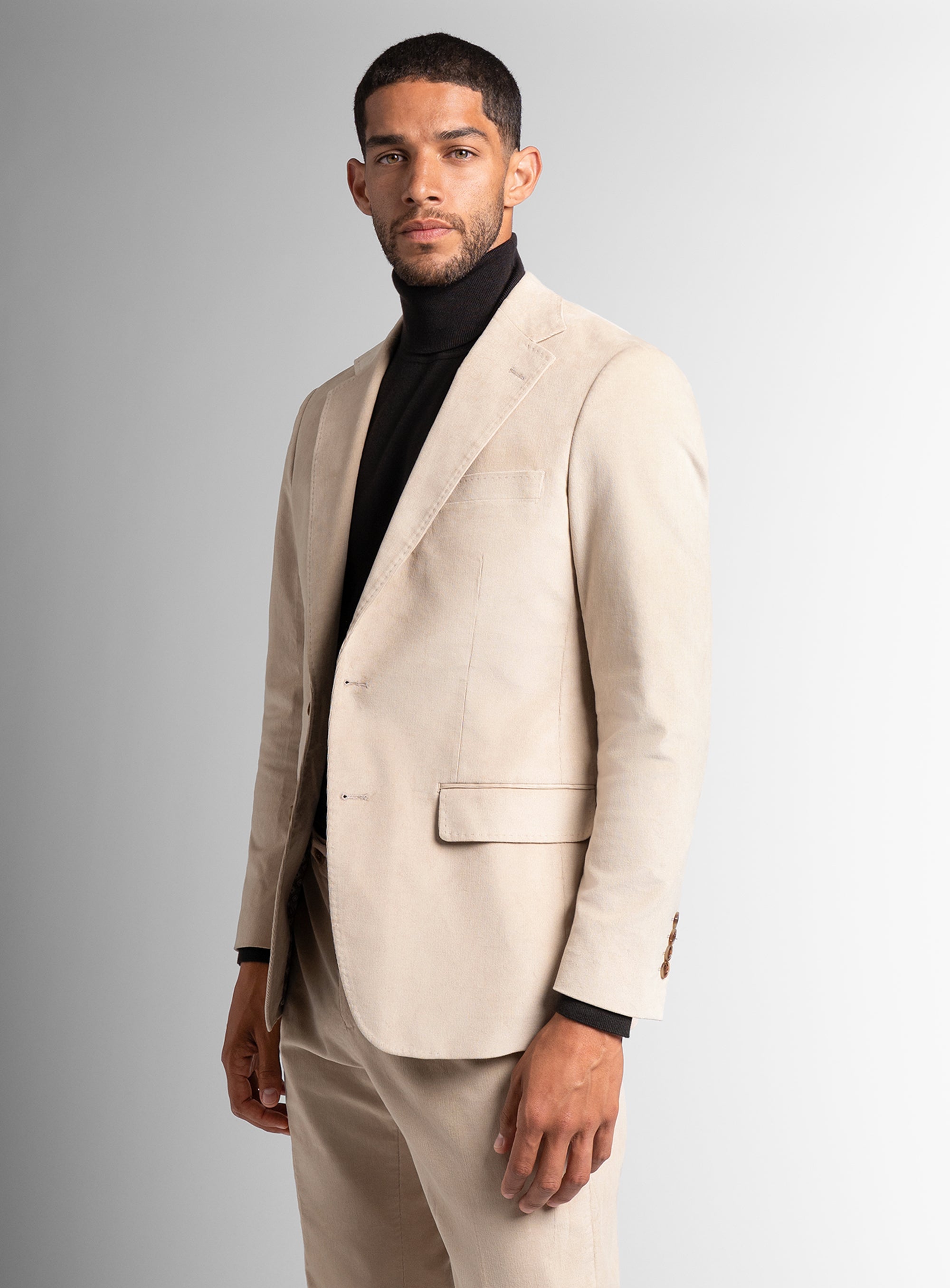 Man wearing a beige suit with a black turtleneck against a gray background
