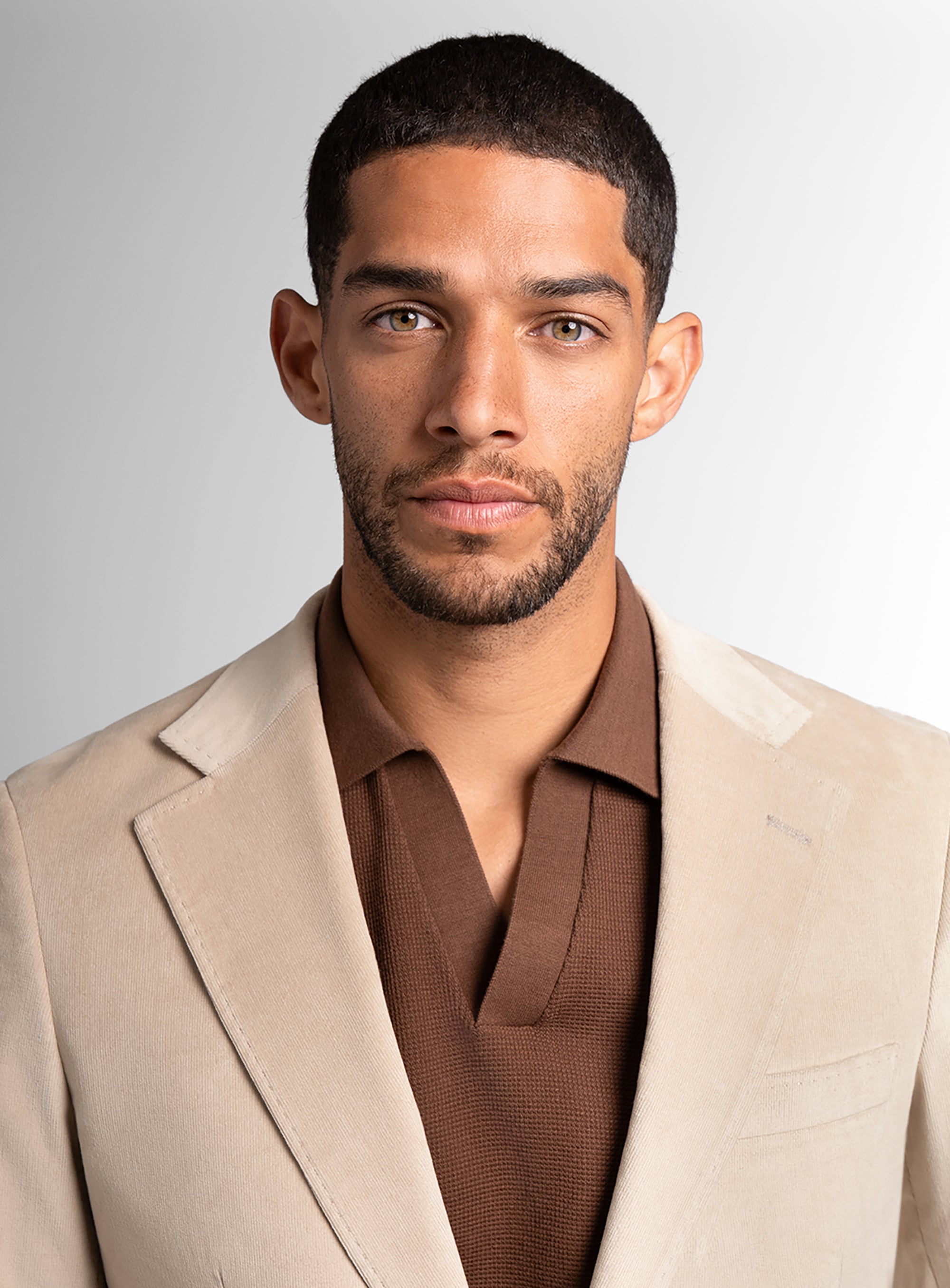 Man wearing a beige blazer over a brown shirt against a gray background