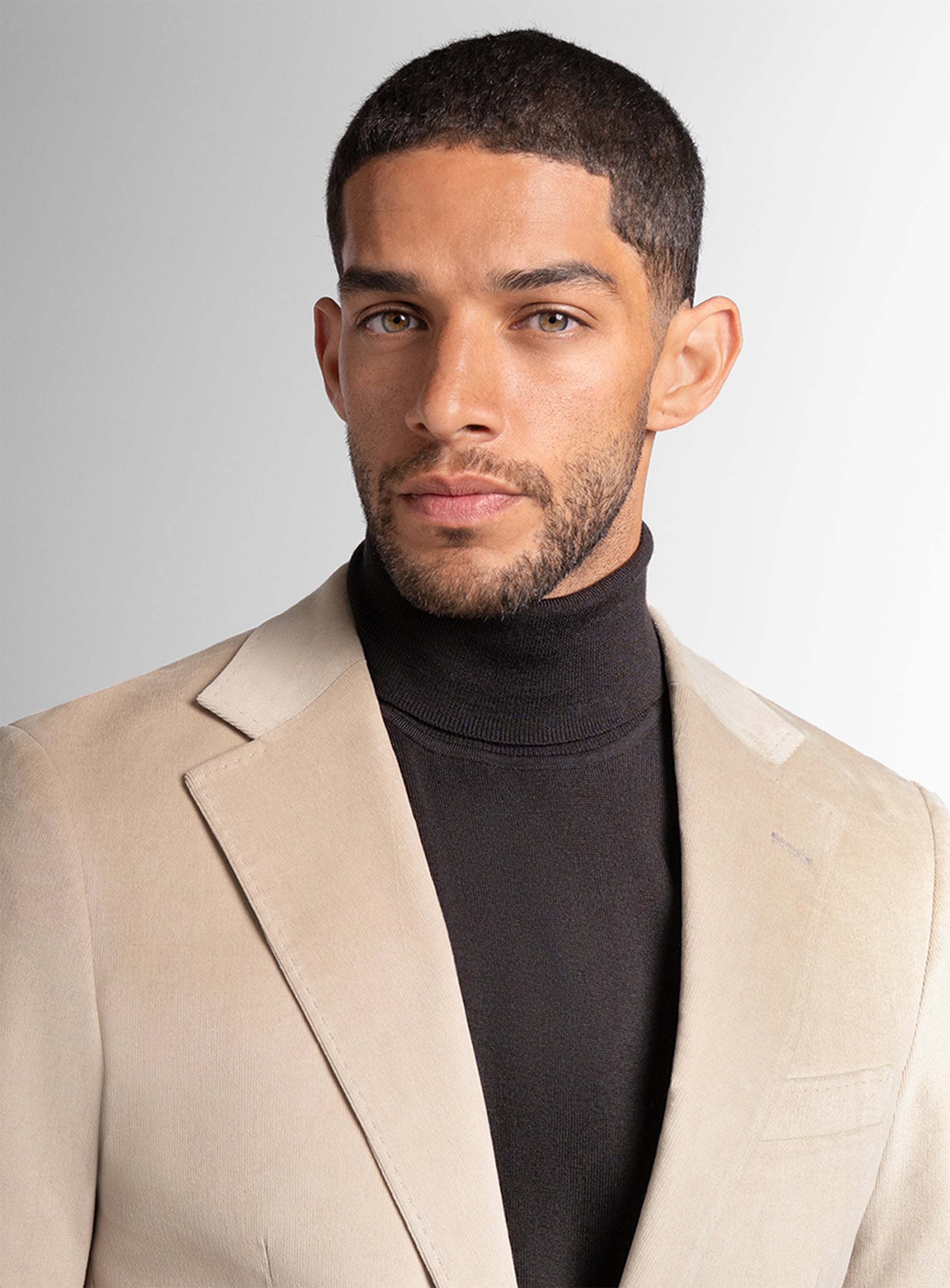 Man wearing a beige blazer over a black turtleneck against a plain background