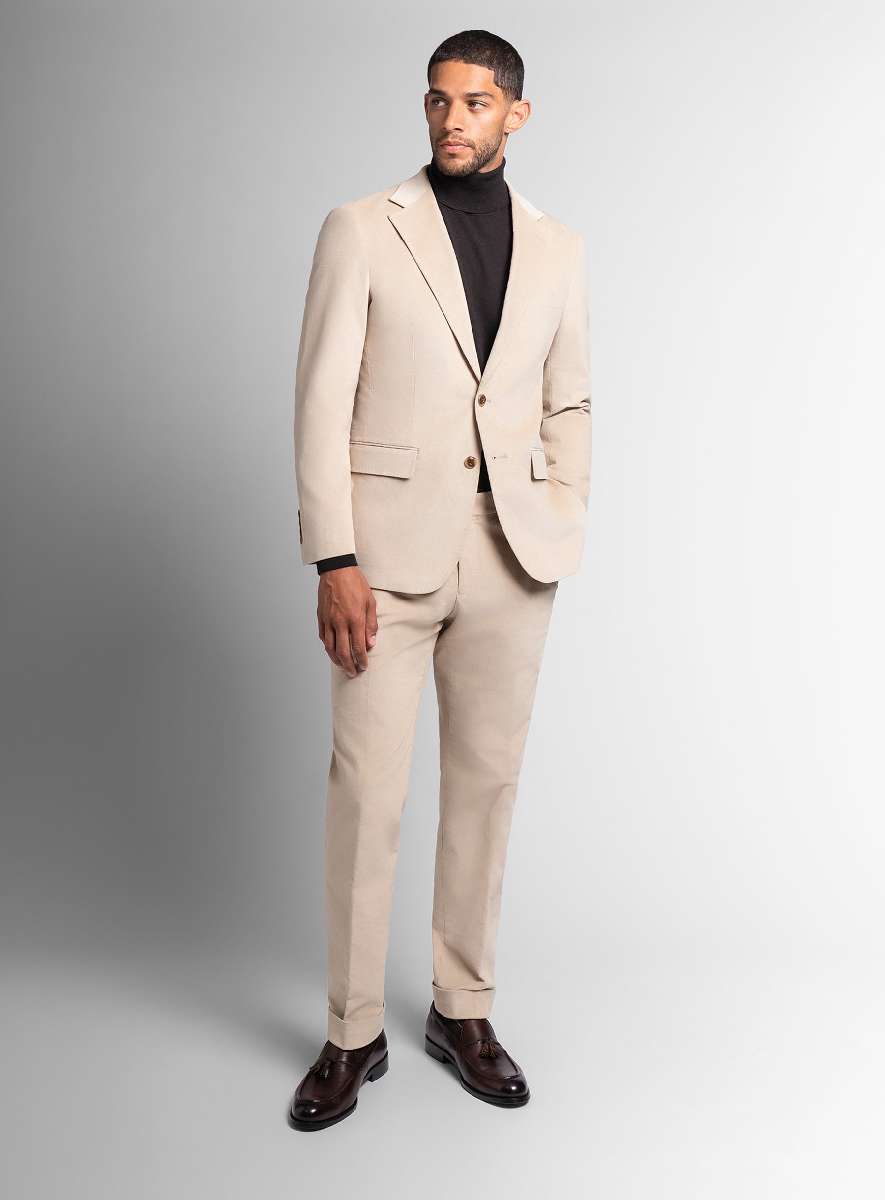 Man wearing a beige suit with a black turtleneck and brown shoes on a gray background
