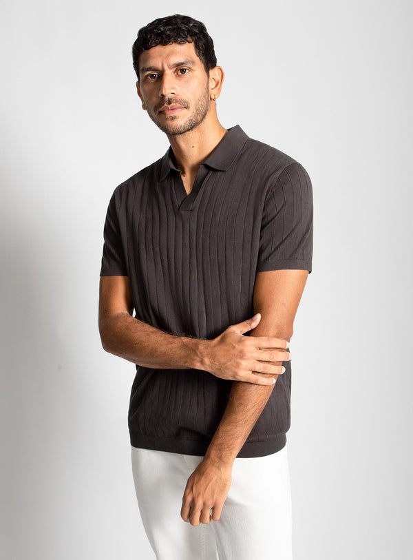 Man wearing a dark brown ribbed polo shirt and white pants on a light gray background