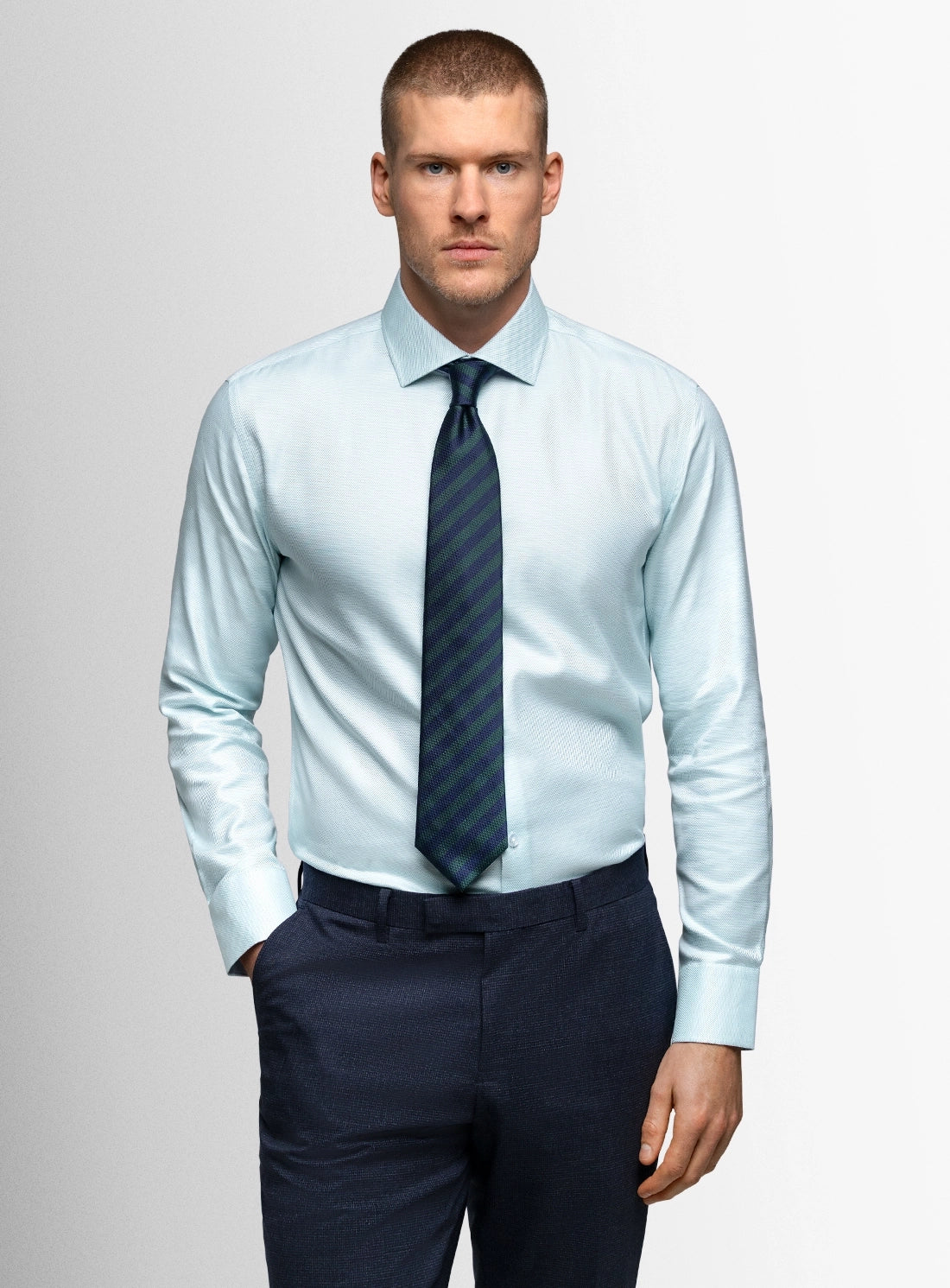 Man wearing a light blue dress shirt, dark tie, and navy pants against a gray background