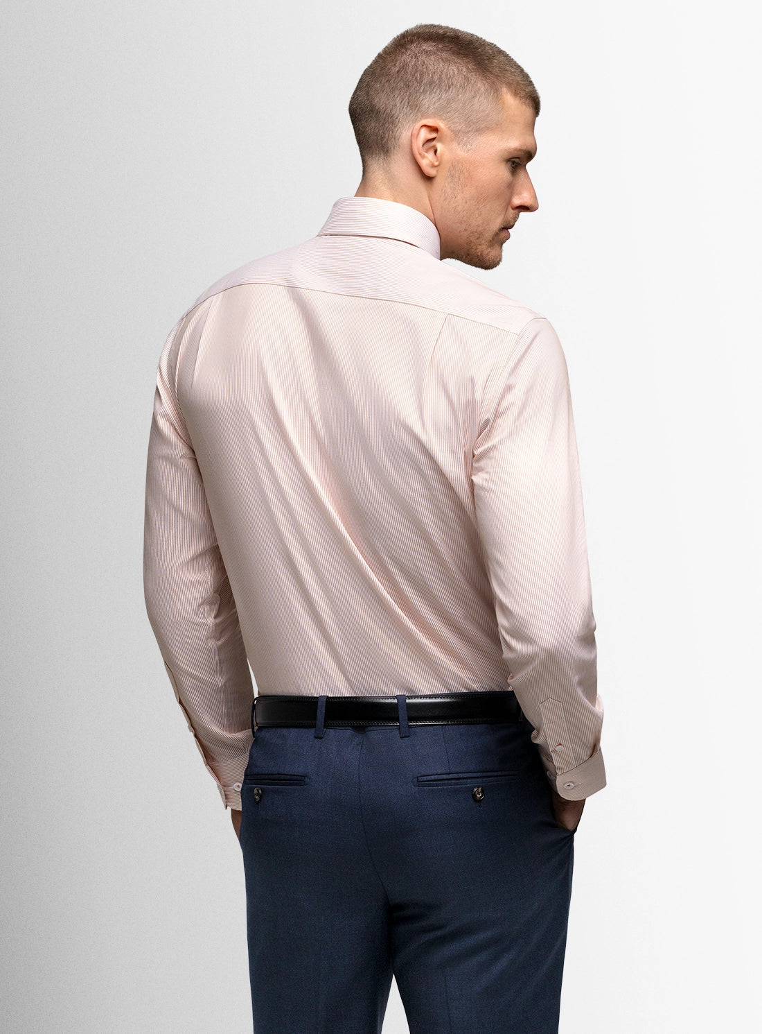 Man wearing a light pink dress shirt and dark blue pants against a plain background