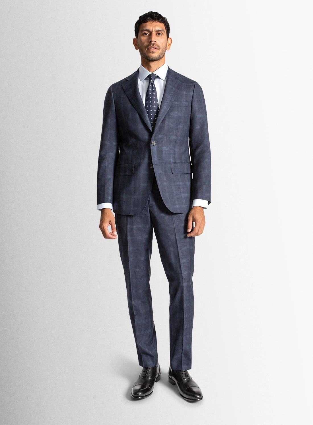 Man wearing a blue checkered suit with a white shirt and black shoes on a light gray background